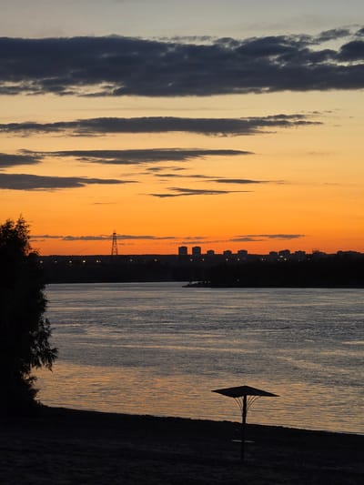 This is a vertical, eye-level shot of a sunset over a body of water, likely a river or lake, with a silhouetted cityscape in the background. The sky is painted with vibrant orange and yellow hues, transitioning to a lighter gray at the top, with streaks of dark, moody clouds. On the right side of the frame, the water reflects the warm colors of the sunset, creating a shimmering, textured surface. A solitary, dark object resembling a satellite dish on a pole stands prominently in the water, silhouetted against the water's surface.

In the distance, the dark silhouette of a city is visible along the horizon, punctuated by small, scattered lights. A tall, lattice-like structure, possibly a pylon or tower, also stands silhouetted against the bright sky. The bottom of the frame shows the dark, dense foliage of trees and bushes, further emphasizing the silhouettes against the fading light.

The time of day is clearly sunset, indicated by the dramatic colors in the sky and the long shadows. The weather appears clear with some cloud cover. The presence of a cityscape suggests an urban or semi-urban environment. The overall mood is tranquil and picturesque, capturing the serene beauty of a sunset. There is no visible text in the image.