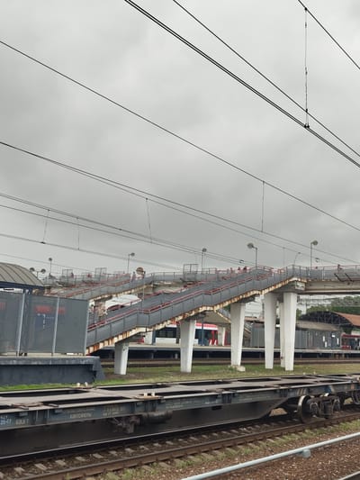 This is a photograph taken from a low angle, looking upwards towards a pedestrian overpass at a train station. The sky is overcast, with a thick layer of grey clouds. Multiple black electrical wires stretch across the upper portion of the frame.

The overpass is constructed of concrete pillars and a metal structure with stairs and ramps. It has red railings and appears to be somewhat aged, with some rust visible. Several people are seen walking on the overpass. Some are walking up the stairs, while others are on the walkway.

Below the overpass, train tracks are visible, with a flatbed railway car positioned on the tracks in the foreground. This car has some Cyrillic text printed on its side, indicating it is for transporting containers. In the background, several other train cars or structures are partially visible, along with a canopy or roofed area. The environment appears to be a functional, active train station. The text on the flatbed car identifies the location as Ramenskoye, Russia.
