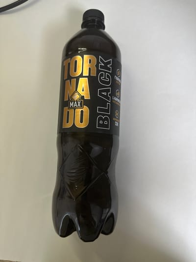 The media file shows a black plastic bottle of "TORNADO MAX BLACK" energy drink. The bottle is lying horizontally against a white background. The label is predominantly black with gold lettering. The text "TORNADO MAX BLACK" is clearly visible, with "TORNADO" in large gold letters stacked vertically on the left, "MAX" in smaller gold letters in the center, and "BLACK" in large gold letters horizontally across the middle of the bottle. There are smaller text elements on the label, including "ГУАРАНА ЭНЕРГИЯ", "L-КАРНИТИН ВООСТАНОВЛЕНИЕ", and "НАТУРАЛЬНЫЙ СОК". The bottle has a textured design on its right side, featuring diamond-shaped patterns and what appears to be a stylized depiction of a tornado. The bottle has a black cap. The lighting suggests it is daytime, and there are no other objects or people visible in the frame. The image is taken in Tambov, Russia.
