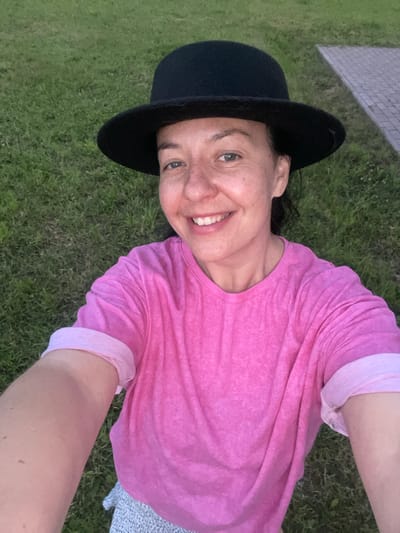 A selfie features a woman wearing a black fedora and a bright pink t-shirt. She is smiling, with her eyes looking towards the camera. The background is predominantly green grass, with a section of grey paving stones visible to the right. There are no discernible landmarks that would indicate a specific location within Timmolovo, Russia. The lighting suggests it is either early morning or late evening.
