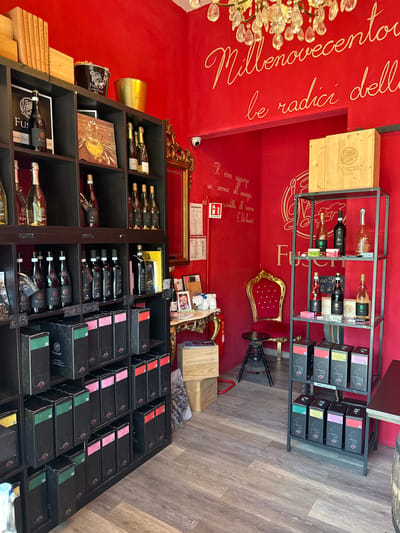 The image depicts the interior of a shop, likely a wine shop, located in Silvi, Italy. The walls are painted a vibrant red and feature gold calligraphy with Italian text. Prominently displayed are shelves filled with bottles of wine and boxed wine sets. On the top shelf, there are books, two metallic buckets, and several bottles. The shelves are made of dark wood. To the right of the shelves, a metal rack displays various bottles and boxed items. In the foreground, more boxed items are arranged. A red velvet chair with ornate gold detailing sits in the middle ground, next to a small round stool. A security camera is visible on the upper right wall. The floor is covered with wood-effect tiles.