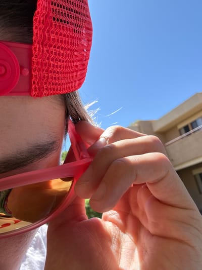 A close-up shot from below shows a person holding pink sunglasses in their right hand, positioning them over their forehead. The person has brown eyebrows and fine, blonde baby hairs peeking out from under a red mesh baseball cap with matching red headphones attached. A building is visible in the background, partially obscured by the hand and sunglasses. The sky is blue. The location is believed to be in Indian Wells, United States of America.