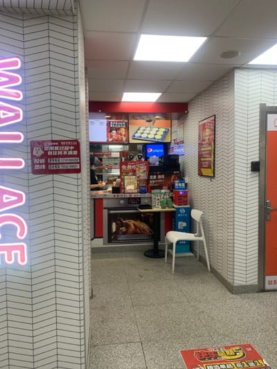 The image depicts the brightly lit interior of a Wallace fast-food restaurant, identified by a partial pink neon sign on the left, located in Shenyang City, China.

The environment features white walls with a repeating dark gray chevron pattern and a gray speckled tile floor. The ceiling is a dropped tile design with multiple square fluorescent light panels providing illumination.

On the left, a vertical pink neon sign partially displays "WALLACE." Below it, a red and white sign with Chinese text and a chicken mascot, likely a customer service notice, is affixed to the wall.

In the background, a service counter with red and white elements is visible. Behind the counter, a person, possibly an employee, is partially seen from the waist up, facing away from the camera, engaged in tasks. Above the counter, multiple digital displays show menu items, including a tray of egg tarts and promotional graphics featuring stylized figures. A Pepsi logo is also visible on one screen. Below the counter, a large graphic depicting an oven with a roasted chicken inside is displayed. Various fast-food items, dispensers, and promotional materials are arranged on the counter.

To the right of the counter, a small, dark rectangular table with a central base stands. Adjacent to it, a white plastic chair is positioned against the chevron-patterned wall. A blue and white stand, likely a charging station, is situated near the chair. Further right, a partial orange door and doorframe are visible. On the floor in the foreground, a red and white decal with Chinese characters is present.