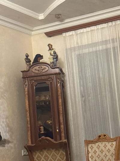 The image shows an interior scene with ornate furniture. The primary object is a dark wood cabinet with decorative carvings and a glass front, revealing some items inside, possibly bottles. On top of the cabinet, there are two figurines, one appearing to be a historical or religious figure with a golden halo and draped clothing. The cabinet is positioned against a wall with a light-colored textured finish. Above the cabinet, a curtain with a subtle striped pattern is visible, suggesting a window or an alcove. The overall aesthetic is opulent, with intricate details on the furniture and figurines. The lighting appears to be artificial, casting a soft glow on the scene. No specific activity or event is taking place; it is a static depiction of the room's decor. The image does not contain any visible text. The setting suggests a well-decorated living space or dining area.