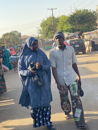 Two individuals are the focal point of this outdoor snapshot. The person in the foreground wears a blue hijab-like garment with a pattern and carries a black bag. Behind them, a younger person sports a checkered shirt, a Nike beanie, and unique lower garments covered in drawings and stickers. They are positioned on a road, likely a bustling area, as evidenced by the surrounding vehicles and pedestrians. The backdrop features buildings, utility poles, and vegetation, suggesting an urban or semi-urban setting.
