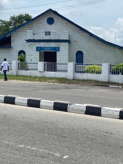 The image shows the Calabar Lodge No. 3416 E.C. Masonic lodge, a white building with blue trim, in Calabar, Nigeria. A man in a white shirt and jeans walks past the building. A black and white curb separates the building from the gray asphalt street. The sky is visible in the background.