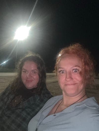 Two individuals are photographed outdoors at night, illuminated by a bright overhead light source. The person on the left has dark, curly hair and is wearing a dark shirt and a plaid jacket. The person on the right has reddish-orange, curly hair and is wearing a light blue collared shirt with a necklace. The background appears to be an open, unpaved area, possibly a parking lot or field, with indistinct structures or poles visible in the distance. The overall scene is dimly lit, with the bright light creating a strong glare and lens flare effect. The location is identified as Kutlu-Bukash, Russia.