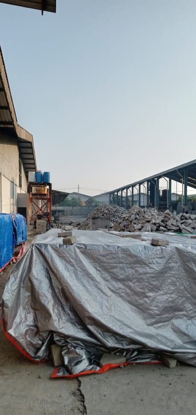 The image depicts an industrial site located in Sumput, Indonesia. In the foreground, a large silver tarp covers an indeterminate pile, secured by a few gray bricks. To the left, part of a blue-covered object is visible near a building's wall, which houses three blue barrels on a rusty metal platform. In the background, a heap of fragmented materials rests next to a long, open-sided warehouse structure. Beyond the warehouse, additional structures are faintly visible in the distance under a clear sky. There are no people visible in the scene.
