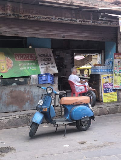 This is a photograph taken in Jodhpur, India, on a street. In the foreground, a blue Vespa-style scooter is parked, with a brown seat and license plate that reads "RJ 01 MY 6294". To the left of the scooter is a weathered metal counter. Behind the counter, a sign is visible with the brand "DODLA" and text in Hindi.
A man, appearing to be in his late 60s or 70s, sits on a red plastic chair to the right of the scooter, facing away from the camera. He is wearing a white shirt and white pants. The background shows a shop with its shutter partially open, revealing a dark interior. A stack of bottled water is visible on a shelf to the right of the man. A yellow sign with Hindi text, advertising "Chapati Centre", is also on the right. The ground is paved with stone slabs, and there are some debris and stains on the pavement. The shop awning above the shutter has Hindi text painted on it. The overall scene depicts a typical street-side shop in Jodhpur.