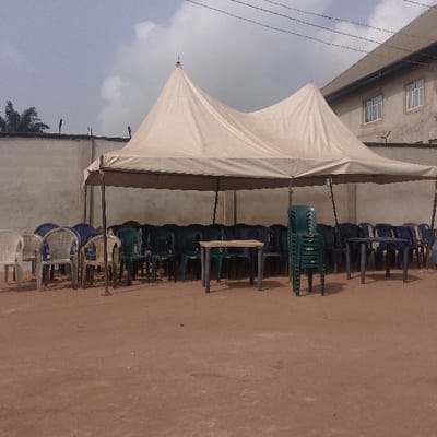 A white tent covers rows of blue, green, and white plastic chairs, along with small tables. The chairs are arranged beneath the tent's shelter on a reddish-brown, dirt ground. A stack of green chairs stands towards the front. Behind the tent, there is a concrete wall on the left and a gray building with windows on the right. Overhead, power lines stretch across a cloudy sky.