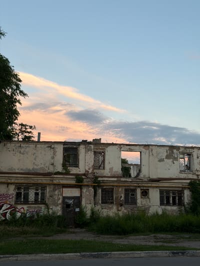The image shows a dilapidated two-story building in Semiluki, Russia, with its roof and upper floor largely destroyed. The facade of the remaining structure is weathered and peeling, with visible signs of decay. Several windows are broken or missing, with some retaining their metal grilles. Greenery, including vines and overgrown bushes, encroaches on the building. Graffiti is present on some parts of the facade and on a closed door. The sky above is a pale blue with wispy clouds tinged with pink and orange from what appears to be a sunset. The foreground shows a patch of grass and a curb bordering a paved surface, possibly a road. The scene conveys a sense of abandonment and disrepair.