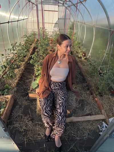 A full shot of a woman with her hair pulled back and wearing a brown button-up shirt and zebra-striped pants sits in a greenhouse. She is looking to the right. Tomato plants are visible in the background, growing in raised beds covered with straw. The greenhouse has a curved, transparent roof supported by metal poles. The ground inside the greenhouse is also covered with straw.