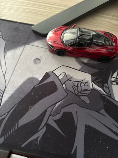A red toy car, possibly a McLaren 720S, is lying on a black and white mousepad with an anime-style design. The mousepad depicts a person wearing a black jacket, with only their arm and shoulder visible. The image was taken in the city of Șelimbăr, Romania. The mousepad is on a wooden surface, with the upper right corner of a black object, possibly a computer monitor, peeking into the frame. The scene is illuminated with overhead lighting. The location is confirmed using the coordinates (latitude: 45.78, longitude: 24.176).