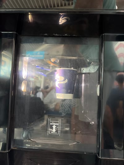 The image shows a close-up of a beverage dispenser. A paper cup with a purple logo is visible inside the dispenser. The cup has the word "BEL" printed on it in small black text. There are lights inside the dispenser that illuminate the cup. A sticker with the word "AUTOMATIC" and upward and downward arrows is on the exterior of the dispenser. The dispenser appears to be in a public indoor setting. The location in the city of Golubovci, Montenegro, is not depicted in the image, nor is it suggested by any elements within the frame.