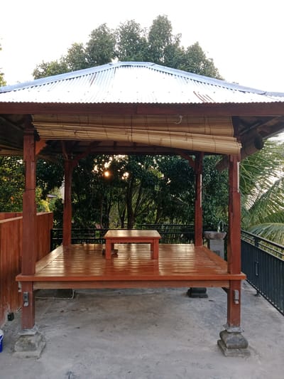 This is a medium shot, eye-level, outdoor photograph of a wooden gazebo situated on a concrete patio. The gazebo has a corrugated metal roof and is supported by four thick wooden posts, each resting on a decorative stone base. The floor of the gazebo is made of polished wood, and a small, low wooden table is placed in the center. A bamboo roll-up blind is partially lowered over the opening on the right side of the gazebo. The background consists of lush green trees and foliage, with the sun's rays filtering through the leaves, indicating it is either sunrise or sunset. In the distance, a black metal railing is visible. The overall scene appears to be a serene and tranquil outdoor space, possibly in a residential or resort setting, within Sawan, Indonesia. There are no people or animals present in the image.