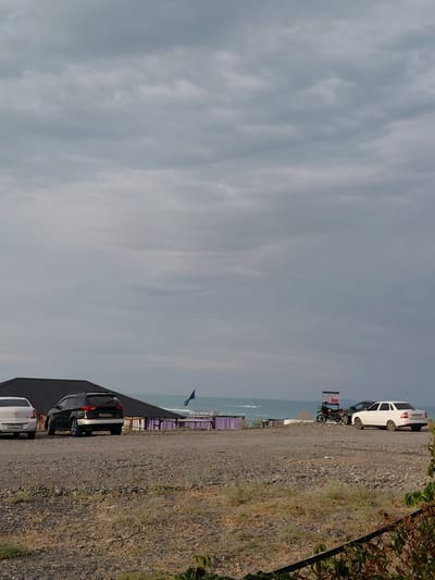 The image captures a cloudy day scene by the sea, likely in Ақши, Kazakhstan. In the foreground, a gravelly, dry area is visible with some sparse vegetation. A dark SUV is parked on the left side, and a lighter-colored sedan is parked to its right. Behind these vehicles, a building with a dark, sloped roof is partially visible. Further back, a blue flag flutters against the backdrop of the sea. The sea itself appears calm, with a jet ski in the distance. On the right side of the image, two white signs with red accents stand upright. A white sedan is parked next to these signs, and a motorcycle is positioned between the signs and a dark car that is mostly obscured. The sky is overcast with numerous clouds, casting a diffused light over the scene.