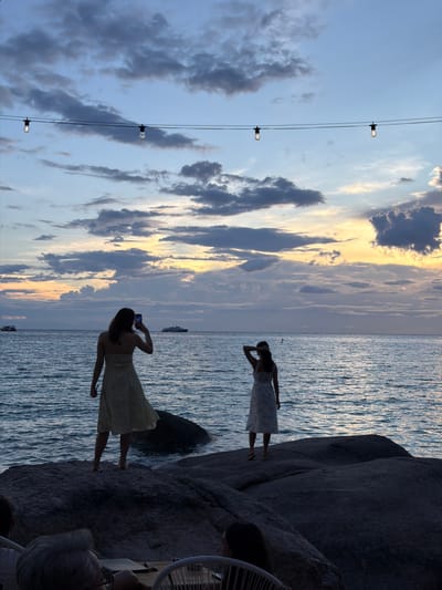 The image depicts a sunset scene at the beach in Ko Tao, Thailand. Three people are visible, two women silhouetted in dresses and one person partially visible from the back in a chair. One woman is standing by the water, looking out towards the sea and holding her hand up to her forehead. The second woman is further back, also by the water, taking a photo with her phone. The sky is filled with clouds, with streaks of orange and yellow from the setting sun. The ocean is calm with ripples reflecting the light. Large rocks are present on the right side of the image, with part of a white chair visible in the foreground, suggesting a seating area. The overall mood is serene and peaceful, with the beauty of the natural landscape as the focal point. There is no visible text in the image.
