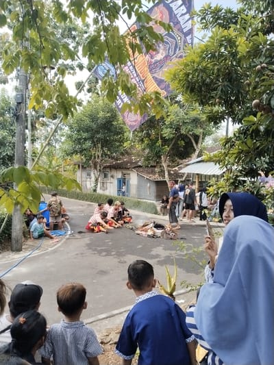 The image depicts a street scene in Mojogedang, Indonesia, where a group of people are gathered. In the foreground, several children are observing an event. To the left, a blue barrel and blue tubing are visible on the side of the road. In the center of the road, a group of individuals in traditional attire are seated on the ground around what appears to be a ceremonial fire or offering. In the background, a modest house with a blue door is situated, surrounded by trees and vegetation. A large banner with a graphic design hangs from a pole, and several fruit-bearing trees are also present. The overall atmosphere suggests a community gathering or cultural event taking place outdoors.