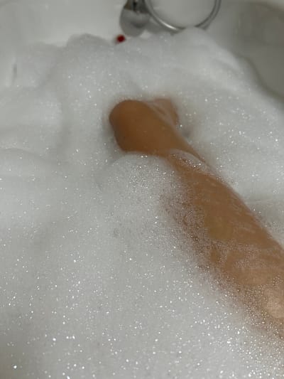 A close-up shot shows a person's leg submerged in a bathtub filled with white foam. The leg appears to be tanned and is partially visible through the bubbles. The foam is abundant and textured, with many small bubbles creating a soft, airy surface. In the upper right portion of the image, a chrome faucet and a small red object are partially visible, suggesting the presence of a bathtub fixture. The overall environment appears to be a bathroom. The location in the city of Vyshevychi, Ukraine, is not depicted or implied by the visual information in the image.