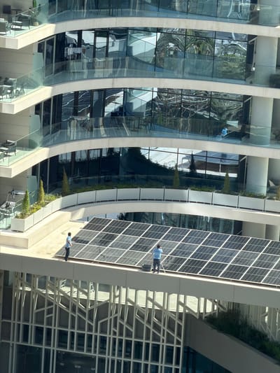 This image captures a high-angle view of a modern, multi-story building facade in Lusail, Qatar, characterized by sleek, curved lines and extensive use of reflective glass. The building's design prominently features sustainable elements, including a large array of photovoltaic solar panels covering a significant portion of its right side.
Several workers, dressed in light blue shirts and dark pants, are engaged in activities along the building's exterior. Two individuals are positioned on a narrow ledge alongside the solar panels; one appears to be standing upright, while the other leans or kneels, possibly performing maintenance or inspection tasks on the panels. Another worker, similarly attired, is visible as a distorted reflection in the highly reflective glass panels on the left, suggesting work on the glass facade itself.
Integrated green spaces are visible along various levels, with lush linear planters providing a natural element against the architectural structure. These planters are positioned on ledges between the glass sections, contributing to the building's aesthetic and possibly its environmental performance. On the far right, a complex, geometric lattice structure in white or light gray adds another layer of architectural detail.
The bright sunlight casts sharp reflections on the glass and illuminates the solar panels, indicating a clear, daytime setting. The scene conveys a sense of ongoing maintenance or construction work on a contemporary, eco-conscious high-rise. No discernible text is visible on the building or the workers' clothing.