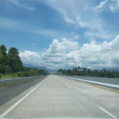 The image captures a view from a vehicle traveling on a highway, likely in or near Seuot Tunong, Indonesia. The highway is paved with concrete and marked with white and yellow lines, indicating multiple lanes. On the left side of the highway, there is a metal guardrail separating the road from a grassy embankment covered in vegetation. Beyond the embankment, a dense forest with tall trees lines the landscape. In the distance, mountains are visible, shrouded in haze, and a few palm trees punctuate the treeline. The sky is a vibrant blue, dotted with scattered white clouds. A pedestrian overpass is visible in the middle distance, spanning the highway. The overall scene depicts a scenic and relatively clear day on a rural highway.