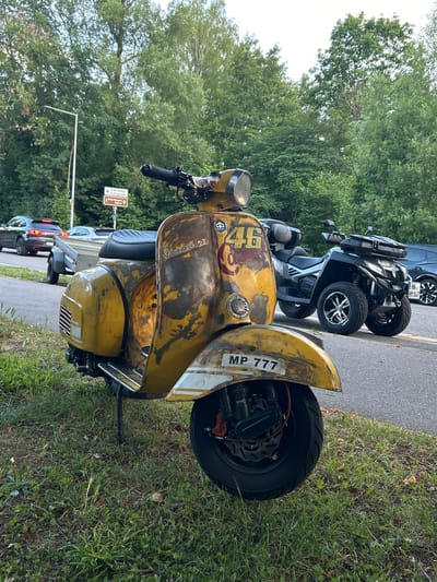 In Heilbronn, Germany, a weathered yellow Vespa scooter is parked on a grassy shoulder beside a road. The scooter has a "46" emblazoned on its side, along with "MP 777" on the front fender. In the background, a black ATV is parked on the asphalt, and two cars are visible on the road, one of which is towing a trailer. Trees line the background, suggesting a semi-rural or park-like setting within the city. The lighting indicates it is daytime, likely late afternoon due to the warm tones.