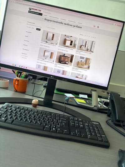 This is a close-up, eye-level, indoor shot of a computer monitor displaying a website for bathroom furniture. The monitor is on a desk, with a keyboard in the foreground. The website is in Bulgarian and features various bathroom cabinets and vanities. The monitor is a black LG brand. The keyboard is a black Logitech brand with a numeric keypad.

The image appears to be taken in an office environment. There are various office supplies visible, including pens in an orange mug, tape, and yellow sticky notes. A black landline telephone is also visible on the right side of the frame. The background is blurred but suggests a typical office setting.

The location is identified as Busmantsi, Bulgaria, based on the context provided, though this information is not visually discernible from the image itself.

There are no people visible in the image. The primary subject is the computer screen displaying the website. The overall scene is static and focused on the digital content on the screen. The lighting is consistent with indoor office lighting.