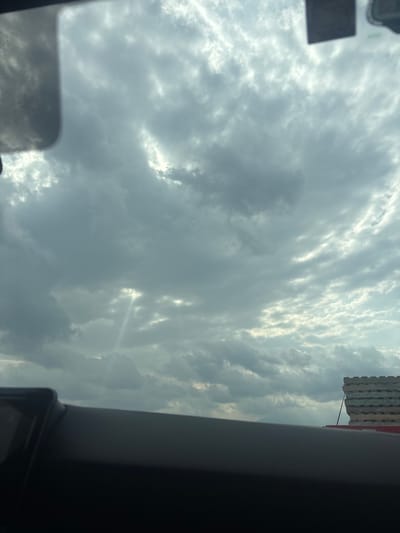 The image shows a cloudy sky, potentially viewed from inside a vehicle. The sky appears to have a mix of light and dark gray clouds, and a few bright patches are visible. Part of a building is visible in the upper right corner. A rearview mirror or other internal vehicle component is in the frame on the left side. The lower right corner shows part of the vehicle interior. The image does not provide evidence to confirm the location in Santa Cruz Tehuixpango, Mexico. There is no evidence about people, actions, or interactions.