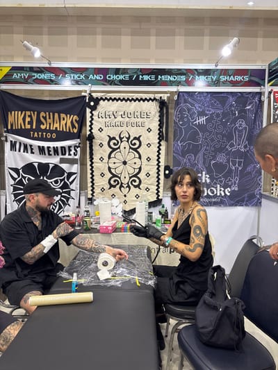 A tattoo booth is depicted within an indoor setting in Ban Rong Ruea Kham, Thailand. Two individuals are prominently featured, engaged with a tattooing station, with a third individual partially visible.
In the foreground, a black tattooing bed is covered with clear plastic sheeting, topped with a roll of white paper towel, a blue spray bottle, and yellow strips. To the left, a male subject with a dark beard and a black baseball cap, wearing a black short-sleeved shirt and black shorts, sits with his heavily tattooed left arm resting on the bed. His forearm is partially wrapped in white material. He is looking downwards at his arm.
To the right, a female subject with short dark hair, wearing a black spaghetti-strap top and black pants, is seated, holding a black tattoo machine in her gloved right hand. Her arms are heavily tattooed, including a large snake design on her right bicep. She is looking directly towards the viewer. On a table behind the bed, various tattoo supplies are arranged, including bottles, a pink box of gloves, and additional paper towels.
On the far right, the back of a third person's head and right shoulder are partially visible. This individual has short, possibly shaved, hair and a gold earring on the right ear, and appears to be observing the scene.
The background consists of three large banners suspended against a light-colored, textured wall. From left to right, the banners display: a black banner with "MIKEY SHARKS TATTOO" and "MIKE MENDES" text above a stylized cat head; an off-white banner with "AMY JONES HAND POKE" and a central black symmetrical design; and a dark blue banner with white outline illustrations and "Stick and Choke" text. Above the banners, a horizontal beam with the text "AMY JONES / STICK AND CHOKE / MIKE MENDES / MIKEY SHARKS" is illuminated by two white spotlights.