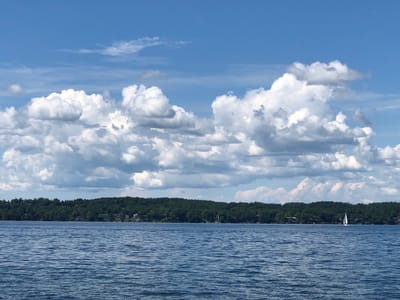 The image depicts a wide, calm body of water, likely Kempenfelt Bay in Barrie, Canada, under a bright blue sky with scattered cumulus clouds. A lush, green treeline with scattered houses lines the distant shore. In the middle of the water, a small white sailboat is visible. The water's surface is textured with ripples, suggesting a gentle breeze.