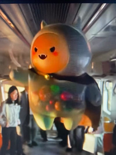 The image depicts a furry, orange-faced character with black eyes, rosy cheeks, and a wide mouth, wearing a grey hood with two small ears. The character has a translucent green body with colorful orbs inside and is holding its left arm out to the side. A small bell is visible around its neck. It appears to be standing inside a public transportation vehicle, possibly a train or bus, with other passengers in the background. The environment shows metal poles and window frames characteristic of a vehicle interior. The scene is not in Beloretsk, Russia.
