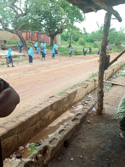 The image depicts a scene in Ilobu, Nigeria (latitude: 7.835, longitude: 4.479). A group of about 15 individuals, primarily women and girls, are walking along a dirt road. They are all wearing blue clothing. The road is lined by a low concrete wall on the right side, with a drainage ditch running along the base of the wall. The ditch contains stagnant water and some debris. The scene is observed from behind a large, weathered wooden post, which is partially visible on the right edge of the frame. The post is situated on the edge of the dirt road. The image is captured from a low angle, providing a partial view of the individuals and their surrounding environment. The overall impression is one of a typical rural scene in Nigeria.