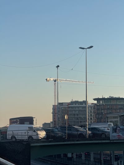 This media file captures a street-level view in Kâğıthane, Turkey, with a focus on urban infrastructure and a construction scene. In the foreground, a metal railing runs along the bottom right, with a dark, textured surface to its left. Beyond the railing, a white van is parked, its side reflecting the sunlight. Adjacent to the van, a large billboard with Turkish text is visible.

Dominating the scene are several tall buildings, some appearing to be residential or office blocks with numerous windows. A prominent construction crane stands tall against a clear, pale blue sky, suggesting daylight hours and fair weather. The crane's structure is intricate, with a long jib and a counterweight at the rear. In the distance, more buildings and another, smaller crane can be seen, indicating ongoing development in the area.

Streetlights with multiple lamps are positioned on poles, one of which is clearly visible in the left half of the frame, reaching towards the sky. The overall atmosphere is one of a busy urban environment undergoing modernization. The lighting suggests it is either early morning or late afternoon, with the sun casting a warm glow on the scene. No people are clearly discernible as subjects in the image, but the presence of vehicles and buildings implies human activity. The image is oriented vertically.