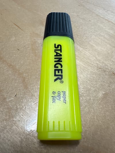 A neon yellow highlighter pen with a black ribbed cap rests on a light brown wooden surface. The brand name "STANGER" is printed vertically in black capital letters on the body of the pen, along with a registered trademark symbol. Below "STANGER", smaller text reads "paper copy & fax" in a similar font. The surface of the wood shows visible grain patterns and a few subtle scratches. The lighting suggests an indoor setting, with shadows cast subtly by the pen. The image appears to be a close-up shot, focusing solely on the highlighter and the surface it is on. There is no indication of a specific location in Lielvārži, Latvia, or any interaction between people and the object.