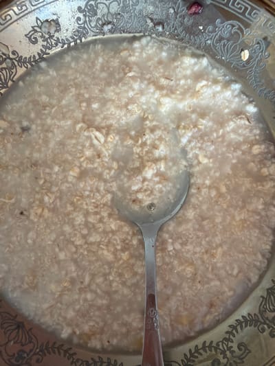 The image shows a close-up, top-down view of a bowl of oatmeal. The bowl is ornate, with a decorative rim featuring intricate floral and geometric patterns. A silver spoon rests in the oatmeal, its handle extending towards the bottom of the frame. The oatmeal itself appears to be cooked with a creamy consistency, with visible oat flakes dispersed throughout. There are a few small, dark specks within the oatmeal, possibly remnants of fruit or spices. The lighting suggests an indoor setting. The location in Lopatino, Russia, is not discernible from the image.