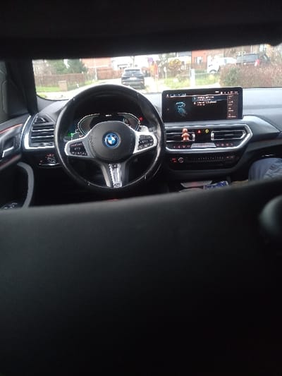 The interior of a BMW is visible, focusing on the steering wheel and dashboard. A large infotainment screen displays navigation information. The steering wheel features the BMW logo. Exterior scenery is seen through the windshield, including a road, buildings, and foliage. The image is taken from the passenger seat.