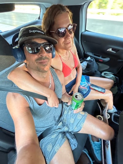 In the interior of a vehicle, likely a car or van, a couple is seated in the back. The male, in the foreground, wears a grey tank top, grey shorts with a pattern, sunglasses, a black "Supreme" baseball cap, and blue headphones. He has his arm around the female, who is seated behind and to his right. She wears a red bikini top and dark blue shorts. She is looking to her right, out the window. The male is holding a green can of what appears to be a beverage and a skateboard is visible next to his leg. Another skateboard is positioned near the female's feet. The interior of the vehicle shows dark upholstery and a seatbelt is fastened across the male. The environment outside the window suggests a sunlit day, possibly a highway or suburban road. The scene appears to be within the municipality of Vidreres, Spain, given the context provided.