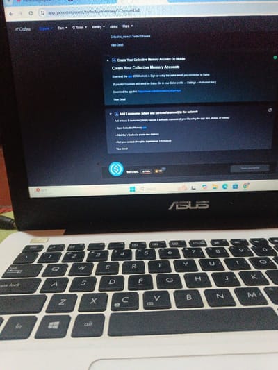 The image shows a laptop computer open to a webpage titled “Create Your Collective Memory Account on Mobile.” The laptop is an Asus model, and the keyboard is black with white lettering. The display is showing a dark background with white text instructing the user to download the app on both Android and Apple phones. The website also states that users must sign up using the same email address connected to their account. There is a green circle in the top right-hand corner of the laptop screen. The laptop is sitting on a white surface with a portion of a red object visible in the background. The location of the image is Purwakarta, Indonesia, with coordinates -6.641 latitude and 107.378 longitude.