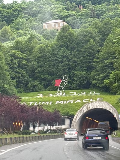 This is an eye-level, medium shot of the Chakvi Miracle Tunnel in Chakvi, Georgia. A large, white, stylized inscription on a grassy hillside reads "CHAKVI MIRACLE" in English, with Georgian text above it. To the left of the inscription, a white outline of a person is depicted kneeling with a red heart. The tunnel entrance, visible in the foreground on the right, is a large, arched concrete structure with lights visible inside, illuminating a truck and two cars driving through. The road leading to the tunnel is wet, suggesting recent rain. Trees and foliage cover the surrounding hills, and a building is partially visible on the crest of a distant hill.