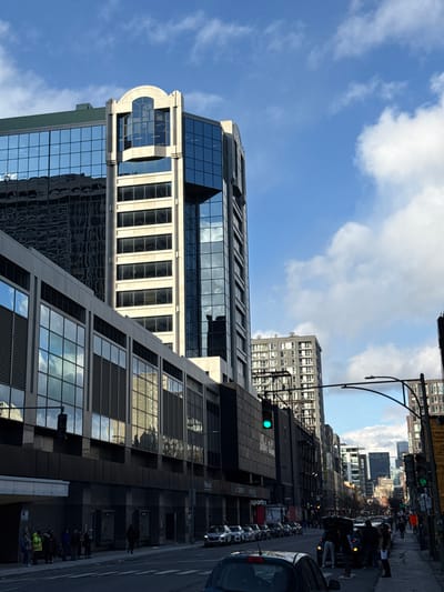 The image depicts a street scene in Westmount, Canada. A modern glass and concrete building dominates the left side, reflecting the blue sky and clouds. Adjacent to it, another building rises, its windows mirroring the surrounding structures. The street is lined with parked cars and moving vehicles, and pedestrians are visible on the sidewalk. Traffic lights hang above the street. A light post with a curved top stands on the right. The sky is blue with scattered white clouds.