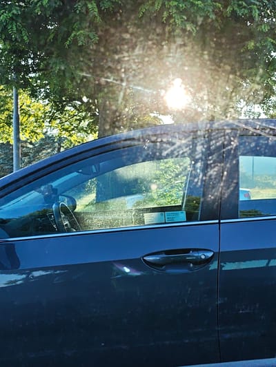 The image shows the side of a dark-colored car parked outdoors, with sun glare and bokeh effects visible on the windows and car body. Trees with green foliage are in the background, along with a street light pole on the left. The car's interior is partially visible, including the steering wheel and rearview mirror. There are stickers on the driver's side rear window, one appearing to be a Toyota emblem. The scene is consistent with a street or parking lot in Mława, Poland, given the typical foliage and street lighting found in the region. There are no people visible in the image.