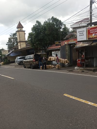 The image displays a roadside scene in Gubbisamamidi, India, under an overcast sky. A gray asphalt road occupies the foreground, bordered by a solid white line and a broken yellow line. On the right side of the road, a brown minivan is parked with its rear hatch open. Two males are engaged near the minivan's rear, handling numerous clear plastic bags filled with light-colored, possibly snack-like, goods; some bags are stacked on the ground. To their right, a third individual, wearing a red shirt, stands beside a wooden frame from which more of these packaged goods are hanging.
In the background, a silver SUV is parked behind the minivan. Further left, a tan-colored mosque minaret with a distinctive rhomboid pattern and a brown roof is visible, partially obscured by trees. A sign reading "Masjid Jami AL-IHYA" is discernible near the mosque structure. A large, dense green tree stands centrally behind the parked vehicles. To the right of the tree, commercial storefronts feature signs, one prominently displaying "BAKSO" and another "WARUNG ANUM AZHAR". On the far right sidewalk, a red and silver fuel dispenser is visible. Overhead, numerous power and communication lines traverse the scene.