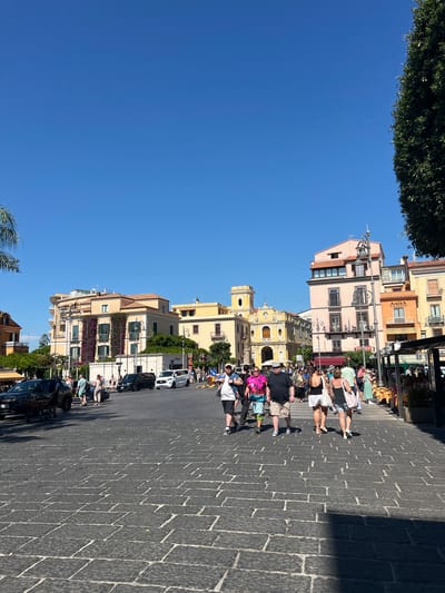 The image depicts a bustling street scene in Sorrento, Italy, under a clear blue sky. The foreground is paved with dark cobblestones, indicating a pedestrian area. A group of people are walking across the street. On the left, a dark car is parked, and further back, a black van and a white van are visible.
The middle ground features a row of buildings with pastel-colored facades, typical of Italian architecture. One building is adorned with climbing purple flowers. A prominent yellow church with a bell tower stands out among the buildings. Several shops and restaurants line the street, with outdoor seating visible.
In the background, more buildings with colorful facades and balconies can be seen. A large green tree is present on the right side of the image, casting a shadow on the cobblestone pavement. The overall impression is of a vibrant and lively urban environment in a Mediterranean city.