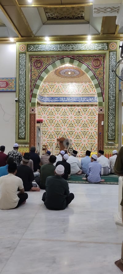 The image depicts a group of men sitting on the floor inside a mosque in Julikeude Dua, Indonesia. They are all facing towards the front of the mosque, with their backs to the camera. The men are dressed in traditional attire, mostly wearing peci hats or sarongs. The mosque features intricate Islamic calligraphy and geometric patterns on its walls and arched entrance. A ceiling fan is visible, suggesting a warm climate. The lighting appears to be artificial, indicating it could be either day or night. The overall atmosphere suggests a religious gathering or prayer session. Text in Arabic script is visible on the walls and archway of the mosque.