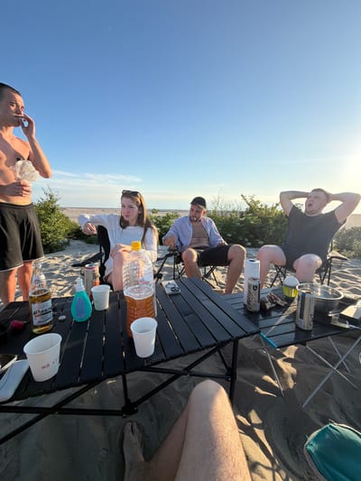 Four individuals are gathered around a black foldable table on a sandy beach. The sky is clear and blue with the sun setting on the right side of the frame. The individuals appear to be casually dressed and are seated in camping chairs. On the table are several drinks, including a bottle of beer, a large plastic bottle filled with a brown liquid, and several cans. There are also white cups and a blue sunscreen bottle. The environment is a sandy beach with some low-lying green vegetation in the background, possibly indicating the outskirts of a settlement or a natural reserve. The scene suggests a relaxed outdoor gathering.