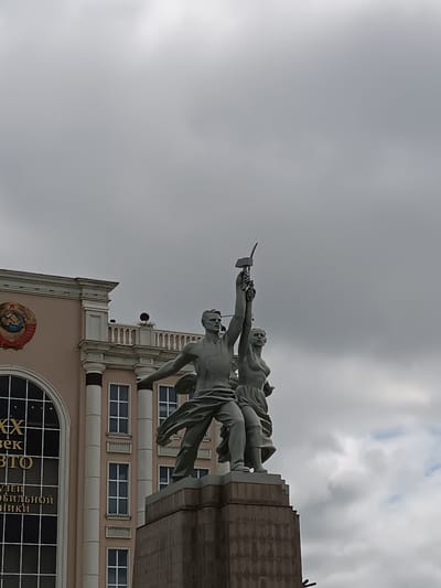 The image shows the Monument "Worker and Kolkhoz Woman" in Verkhnyaya Pyshma, Russia. The monument features a male and female figure, both with outstretched arms, holding a hammer and a sickle respectively, symbolizing Soviet industry and agriculture. The figures are made of metal and stand on a large pedestal. In the background, a light pink building with classical architectural elements and a large arched window with golden lettering is visible. The sky is overcast with gray clouds. The monument is a well-known landmark and a symbol of the Soviet era.