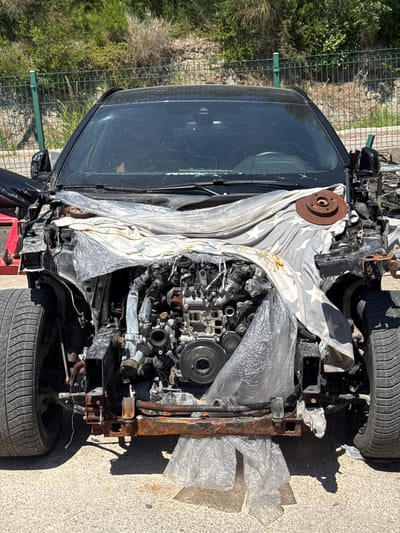 The image shows the front of a heavily damaged black car, likely a passenger vehicle, in an outdoor setting. The car's hood and front bumper are missing, exposing the engine and front-end components, which appear corroded and dirty. A white sheet with a star pattern is draped over a portion of the engine bay, and a rusty brake disc is visible on top of the sheet. The car has two visible tires on the left and right sides. The background includes a green metal fence, some dry vegetation, and a stone wall. The ground surface is asphalt or concrete. The scene is not in a city but rather in a more rural or industrial area, potentially a junkyard or a private property for vehicle disposal. There are no people or animals visible in the image. The lighting suggests it is daytime, likely sunny. The location is identified as Dub, Montenegro.