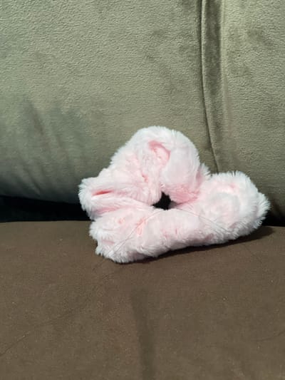 A fuzzy pink scrunchie lies on a brown fabric surface, which appears to be upholstery. Behind the scrunchie, a textured, muted green fabric surface is visible, suggesting the scrunchie is on a couch or chair. The lighting creates soft shadows on the green surface. The image is a close-up, with the scrunchie occupying the center-right portion of the frame. No people or actions are depicted. The location in Bandarbireuen, Indonesia is not determinable from the visual information provided in the image.