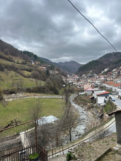 The image presents a panoramic, elevated view of the mountainous village of Erma Reka, Bulgaria, under a heavily overcast sky dominated by dark gray clouds. The village is situated within a valley, flanked by steep, partially forested hillsides.
A shallow, rocky river flows through the center of the valley, winding from the background towards the foreground. On the left bank of the river, a large green field is visible, bordered by a distinct, long, white concrete retaining wall or structure. Above this field, the hillside consists of terraced green areas and numerous bare deciduous trees, with scattered houses featuring light-colored facades and various roof types (red, white) ascending the slope.
On the right bank, a paved road parallels the river, lined with a dense concentration of residential buildings. These houses, predominantly two to three stories, exhibit a mix of architectural styles with varied roof colors including red, orange, and gray, and light-colored exteriors. Several vehicles (cars) are visible parked along this road in the mid-ground. The right hillside also shows a mix of bare deciduous and evergreen trees higher up. Distant mountains are faintly visible in the background through the hazy conditions.
In the immediate foreground, the edge of a concrete structure is visible, featuring a dark metal railing with a lighter-colored top rail and a small, dark green potted plant. To the left, a rustic, gnarled vine-covered structure serves as another barrier. A thick, dark power line extends diagonally from the upper left towards the lower right of the frame, partially obscuring the sky and distant right hillside. A circular speed limit sign (30 km/h) is visible near the road on the right. No individuals are discernible within the frame.