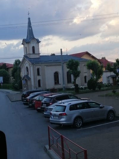 The image shows a street scene in Fălticeni, Romania, with a church prominently featured. The church has a pale yellow facade, a dark blue roof, and a tall, pointed steeple topped with a weather vane. In front of the church, several cars are parked in a row. The foreground shows a silver station wagon. The sky is overcast with some sunlight breaking through the clouds. The overall environment appears to be a quiet residential or town area.